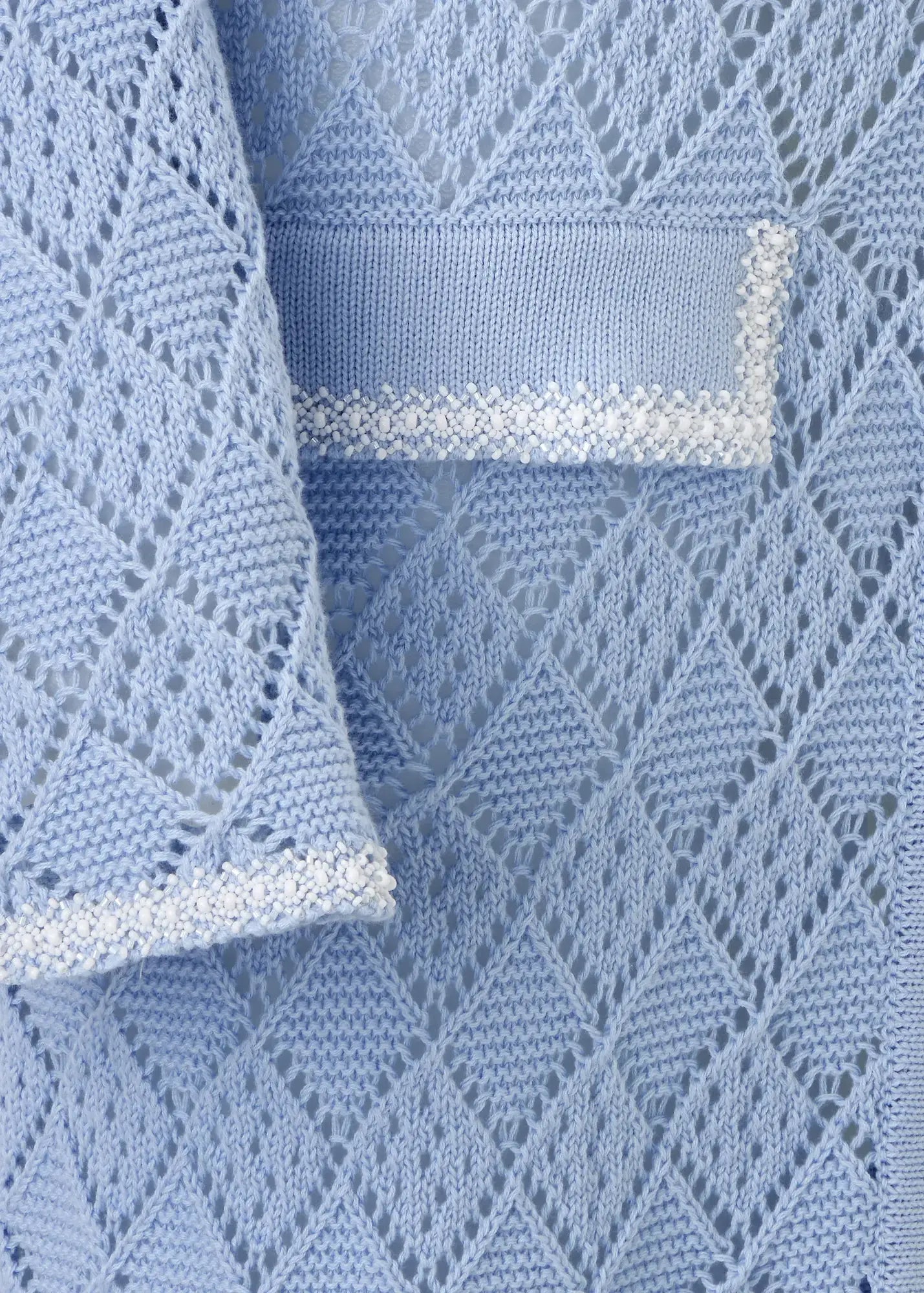 Close-up detail of a luxury sky-blue cotton knit cardigan featuring an intricate openwork geometric pattern, textured trim accents on the pocket and cuff, and exceptional artisanal craftsmanship.