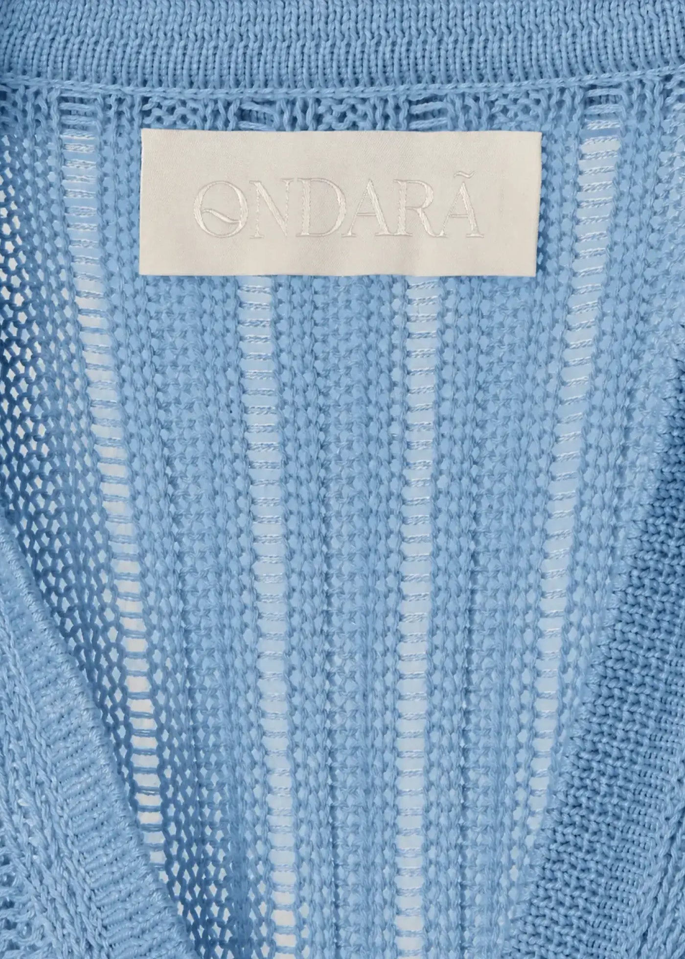 Close-up of the signature label on a luxury designer knit dress made from fine cotton. The embossed Ondara logo is elegantly integrated into a soft woven label, surrounded by delicate open-knit textures and refined craftsmanship.