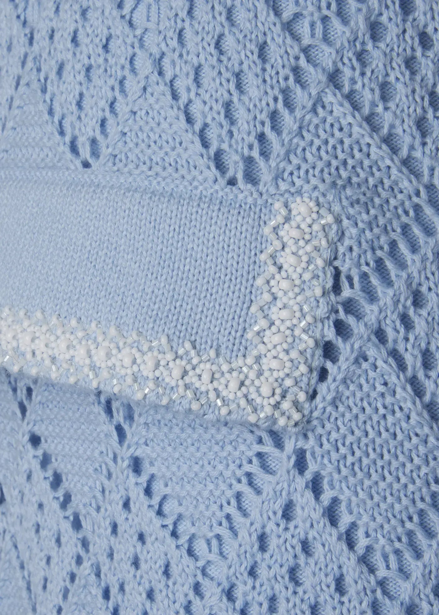 Close-up of luxury sky blue crochet knit shorts featuring a decorative flap pocket embellished with hand-applied white beads. The intricate openwork knit structure and artisanal detailing highlight the refined craftsmanship of this designer Ondara resort wear piece.