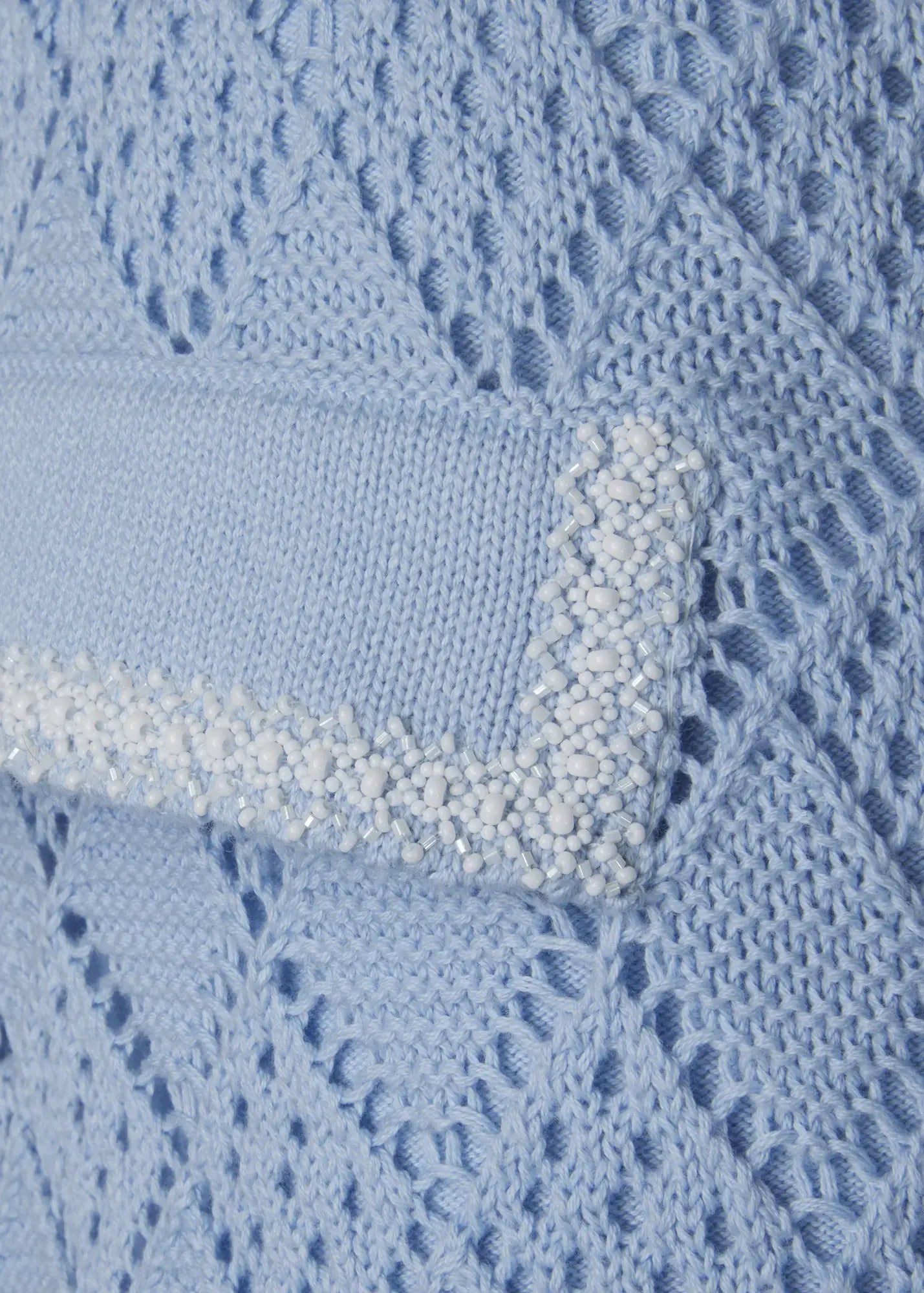 Close-up of luxury sky blue crochet knit shorts featuring a decorative flap pocket embellished with hand-applied white beads. The intricate openwork knit structure and artisanal detailing highlight the refined craftsmanship of this designer Ondara resort wear piece.