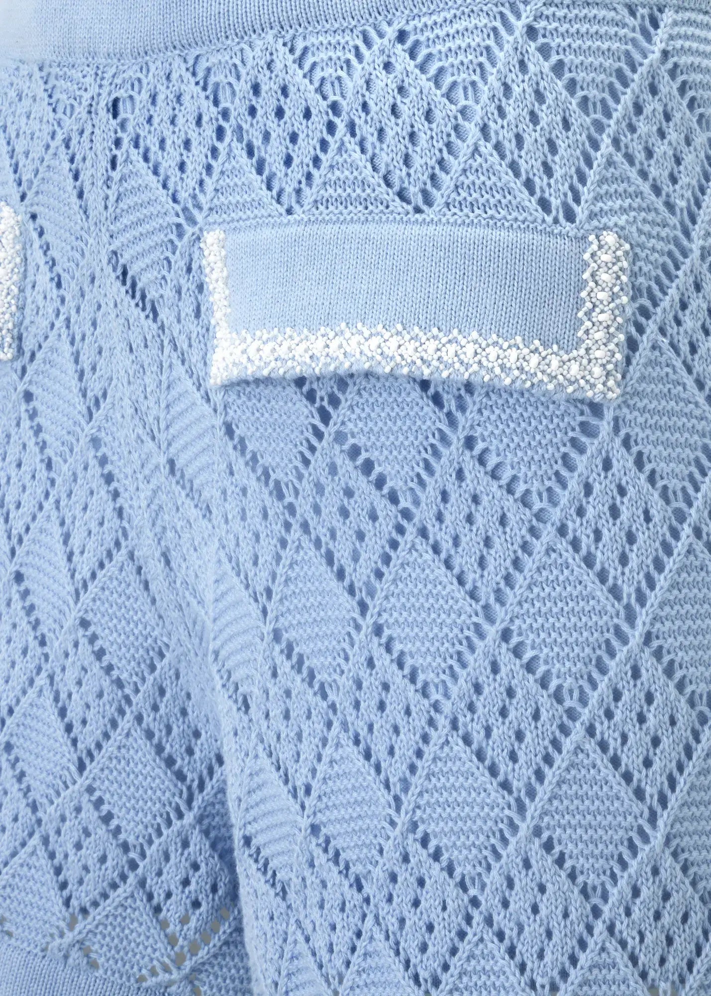 Close-up of luxury sky blue crochet knit shorts featuring an intricate openwork diamond pattern and decorative flap pocket detail adorned with hand-applied white bead embellishments. Crafted from fine cotton knit, this designer Ondara piece combines artisanal craftsmanship with refined resort elegance.